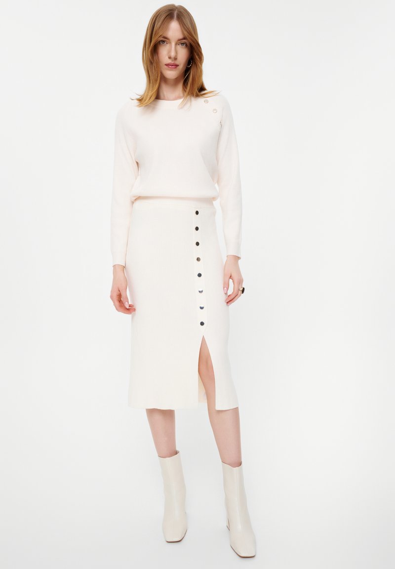 Brede gebreide sweater in lichtbeige met ribbels en knoopdetails, gecombineerd met een crèmekleurige midi-skirt met een zijslit en zwarte knoopaccenten.