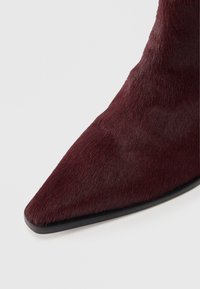 Bottine pointue bordeaux avec une finition douce et texturée ressemblant à de la fourrure et une semelle noire élégante, présentant une silhouette moderne et acérée.