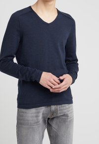 T-shirt à manches longues bleu marine avec encolure en V, en tissu doux et texturé. Présente des manches raglan et un accent subtil à l'ourlet. Associé à un jean gris clair.