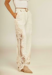 Linnen wijde broek in lichtcrème met ingewikkelde beige kantafwerking langs de zijkanten. Voorzien van zakken en een bijpassende riem met een decoratieve gesp.