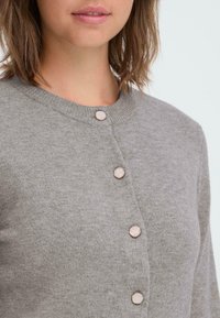 Cardigan gris avec un col rond, doté de cinq boutons en matière contrastante. Tissu tricoté doux et texturé visible en gros plan.