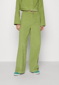 Groene corduroy cropped jas en wijde broek, met een subtiele ribbelstructuur en zilveren knopen, gecombineerd met turquoise schoenen.