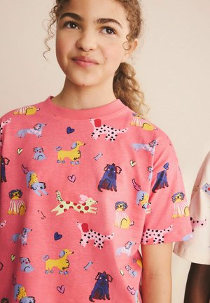 Ung person med krøllet hår iført en lyserød skjorte dækket af farverige tegneseriehund-illustrationer og små hjerter på en beige baggrund.