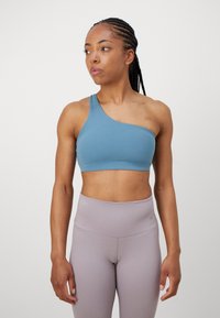 Asymmetrisk sports-bh i blå, laget av stretchy materiale med en glatt tekstur. Kombinert med høyt liv lys lilla leggings.