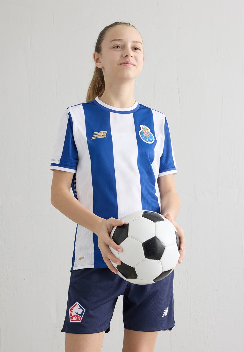 Kék-fehér csíkos focimez logóval, sötétkék rövidnadrággal párosítva, kezében fekete-fehér focilabdával. Semleges háttér.