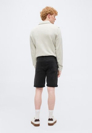 Άνδρας με σγουρά κόκκινα μαλλιά που φοράει ανοιχτό μπεζ πουκάμισο μακρυμάνικο, μαύρο σορτς με γυρισμένα τελειώματα, λευκές κάλτσες και λευκά αθλητικά, γυρισμένος πλάτη σε απλό φόντο.