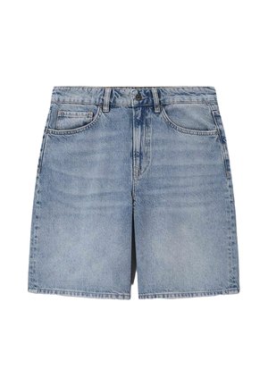 Vrouw staat, draagt gelaagde blauwe spijkerjassen, spijkershorts en bruine geweven sandalen tegen een beige achtergrond.