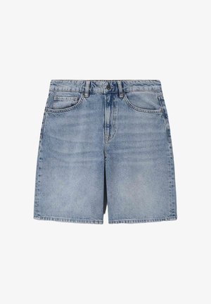 Femme debout portant plusieurs vestes en denim bleu superposées, un short en denim et des sandales tressées marron sur un fond beige.