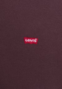 Burgundská tkanina značky Levi's s malým červeným vyšitým logem a bílým textem. Materiál je texturovaná bavlna, která poskytuje klasický vzhled.