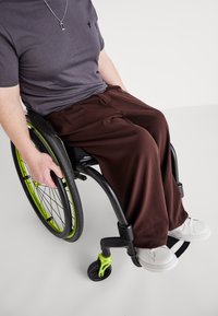 Un pantalon marron drape sur les jambes d'une personne en fauteuil roulant, associé à un T-shirt gris et des chaussures blanches. Le fauteuil roulant présente des accents verts.