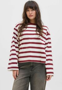 Model in een maroon en wit gestreepte lange mouw shirt, gecombineerd met donkergrijze jeans, staat tegen een effen achtergrond.