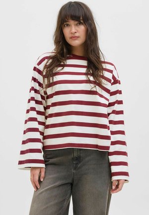 Model in een maroon en wit gestreepte lange mouw shirt, gecombineerd met donkergrijze jeans, staat tegen een effen achtergrond.