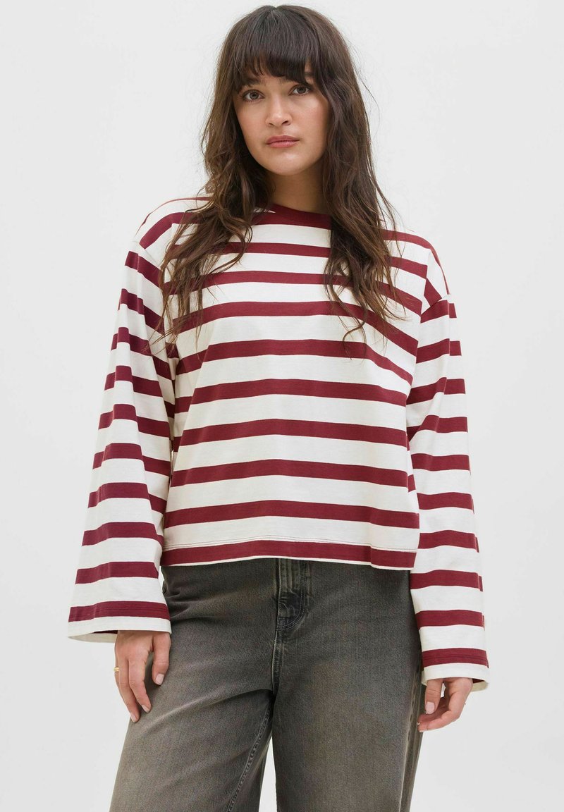 Model in een maroon en wit gestreepte lange mouw shirt, gecombineerd met donkergrijze jeans, staat tegen een effen achtergrond.