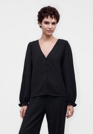 Junge Person mit kurzen lockigen Haaren, die eine schwarze, strukturierte Bluse mit langen, ballonartigen Ärmeln und passende schwarze Hose trägt.