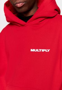 Persona che indossa una felpa rossa con cappuccio, con la scritta bianca "MULTIPLY" sul petto, visibile dalla gola fino alla parte superiore del torso.