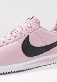 Rosa sportskor med en mjuk tygexteriör, svart swoosh-logotyp och strukturerad vit sula. Snörningen är ljusrosa, vilket ger en kontrast.