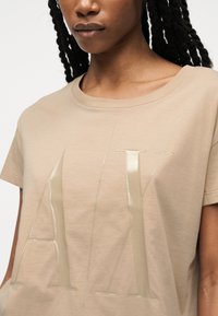 Γυναίκα που φοράει μπεζ μπλουζάκι με κοντά μανίκια και στρογγυλή λαιμόκοψη, με μεγάλα λαμπερά γράμματα "A|X" ανάγλυφα στο μπροστινό μέρος.