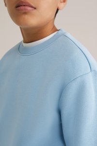 Gaiši zils kreklveida džemperis, izgatavots no mīksta auduma. Iezīmējas ar ribotu apkakli un plecu šuvēm, ar minimāli redzamu šuvju. 