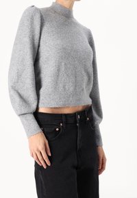 Grijze gebreide trui met hoge hals en pofmouwen, cropped design, gecombineerd met zwarte jeans met koperen accenten en een knoopsluiting.