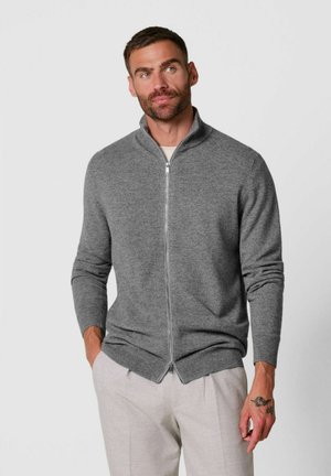 Uomo con capelli corti e barba indossa un maglione grigio con zip e pantaloni chiari, in piedi con le mani parzialmente nelle tasche.