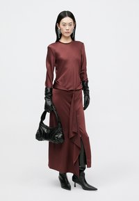 Vestito bordeaux con maniche lunghe e davanti drappeggiato, abbinato a guanti in pelle nera e una borsa nera con texture, completato da tacchi alti.