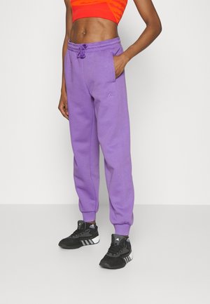 Osoba ubrana w fioletowe spodnie dresowe Adidas, czarne buty sportowe Adidas oraz czerwony krótki top, stojąca na tle jednolitego jasnego tła.