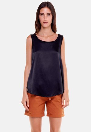 Femme portant un haut noir sans manches et un short orange brûlé à ourlet roulé, debout devant un fond clair uni.