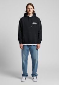 Felpa nera con cappuccio con testo "AMOUR", tasca frontale, abbinata a jeans azzurri chiari e sneakers bianche, con tre strisce nere.