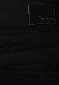 Jean en tissu noir avec une texture tissée, un motif cousu et une étiquette noire de marque avec des lettres blanches.