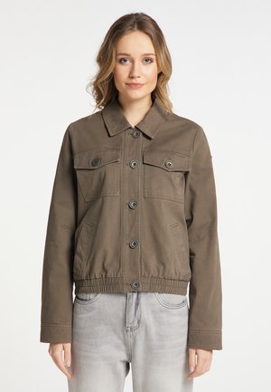 Woman with light brown hair wearing a buttoned brown jacket with chest pockets and light gray jeans, standing against a plain white background.