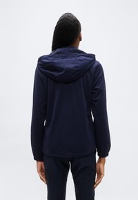 Άτομο με μακριά μαλλιά που φοράει μια μπλε ναυτική κουκούλα και αντίστοιχο παντελόνι, στέκεται με την πλάτη προς τα εμάς μπροστά σε ένα απλό φόντο.