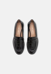 Mocasines de charol negro con textura acolchada y parte superior suave. Diseño sin cordones con un tacón bajo y detalles de costura en los lados.