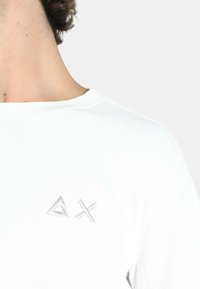 T-shirt bianco in cotone con scollo rotondo e maniche corte, con simboli geometrico ricamati in argento sul lato sinistro del petto.