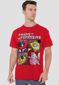 Red cotton t-shirt featuring a graphic with four Transformers characters in comic-style panels; includes vibrant colors and outlined details.