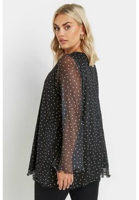 Blusa nera trasparente con pois bianchi, orli scallop e maniche svasate. Taglio morbido, lunghezza media, tessuto leggero.