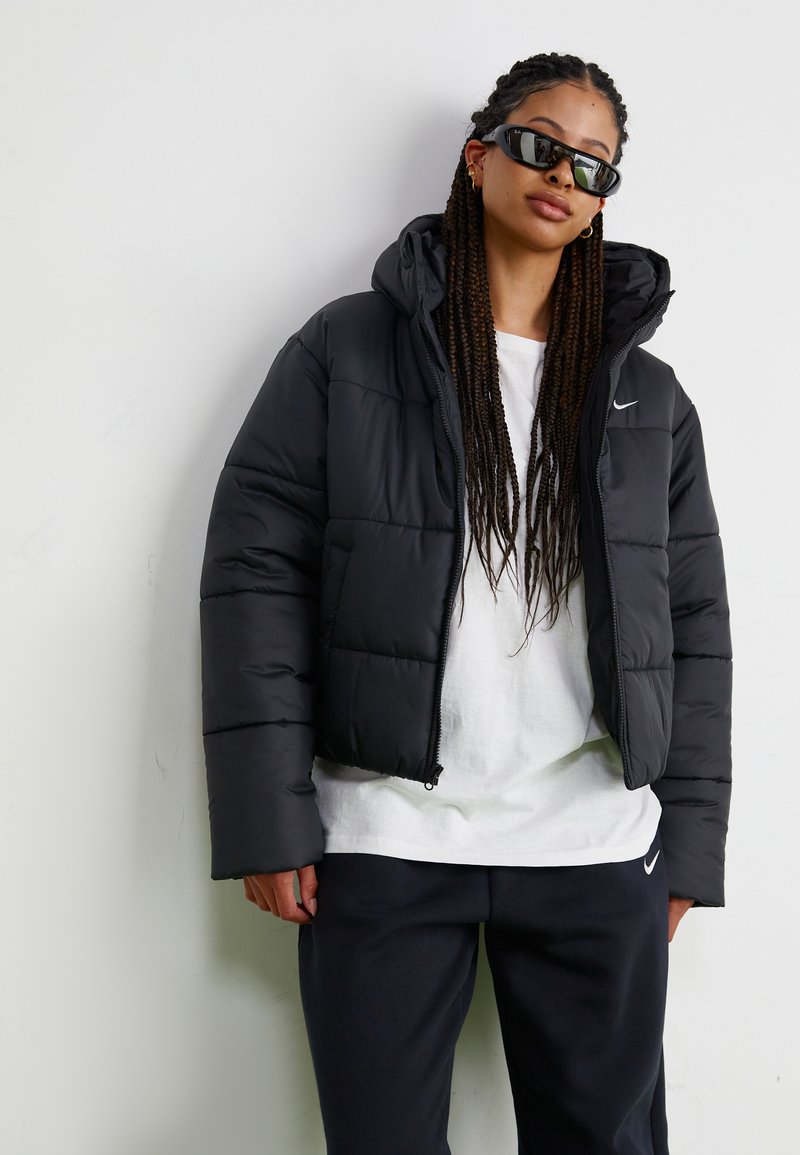 Giubbotto imbottito nero con cappuccio e zip frontale, caratterizzato da cuciture orizzontali e un piccolo logo Nike sul petto. T-shirt bianca sotto.