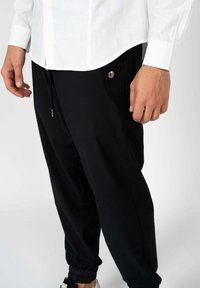 Zwarte joggerbroeken met elastische boordjes, voorzien van een trekkoord in de taille en zijzakken. Een metalen knoopaccent is zichtbaar op de linkerzak.