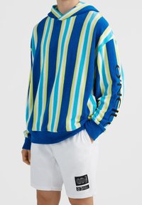 Personne portant un pull à capuche rayé bleu, vert et blanc avec le texte "O'Neill" sur la manche et un short blanc avec un patch logo noir.