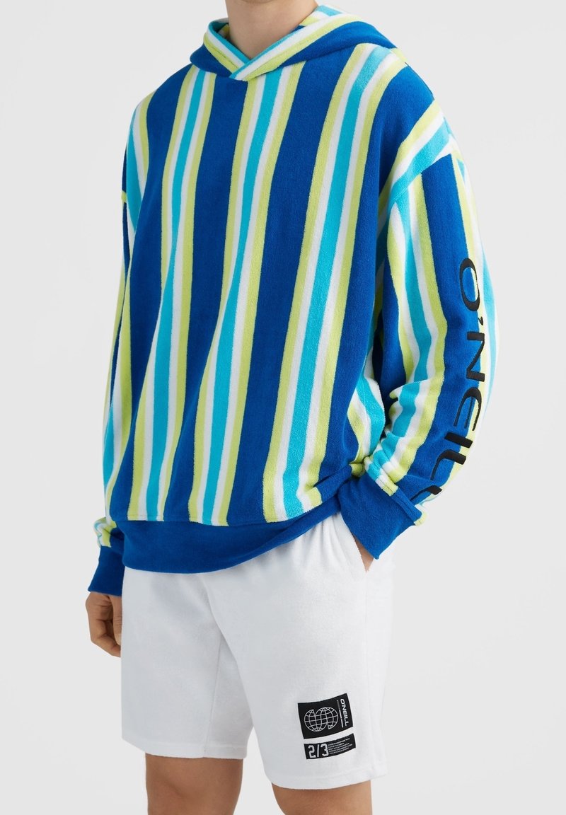 Personne portant un pull à capuche rayé bleu, vert et blanc avec le texte "O'Neill" sur la manche et un short blanc avec un patch logo noir.