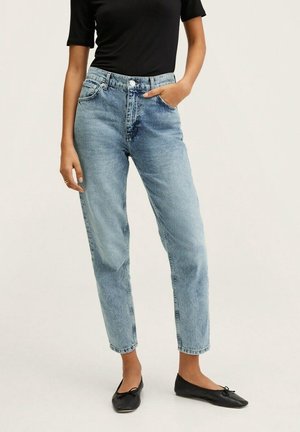 Frau trägt hoch taillierte hellblaue Jeans, schwarzen kurzärmeligen Oberteil und schwarze Ballettschuhe, eine Hand in der Tasche.