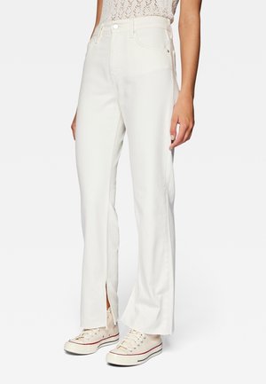 Femme portant un jean blanc taille haute avec des fentes à l'avant des jambes, associée à des baskets beiges et un haut en dentelle blanche, debout devant un fond uni.