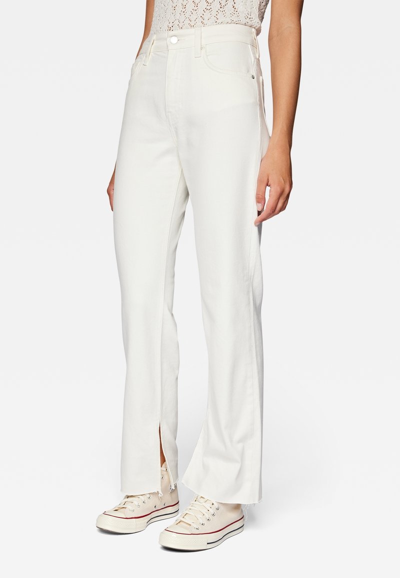 Femme portant un jean blanc taille haute avec des fentes à l'avant des jambes, associée à des baskets beiges et un haut en dentelle blanche, debout devant un fond uni.
