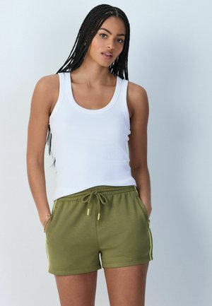 Žena s dlhými vrkočmi, ktorá má na sebe biely tielkový top a olivovo zelené šortky so šnúrkou, stojí s rukami v vreckách.