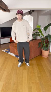 Sweater em tom bege claro com riscas, calças largas pretas e ténis prateados. O fundo apresenta uma planta e um sofá laranja texturizado.