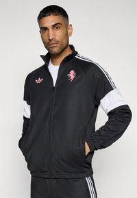 Μαύρο αθλητικό μπουφάν με φερμουάρ, λευκές λεπτομέρειες και ροζ κεντημένο λογότυπο αλόγου. Διαθέτει τσέπες με φερμουάρ και όρθιο γιακά.