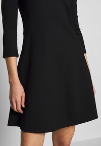 Black fitted dress with three-quarter sleeves, a smooth texture, and flared skirt. Features minimal stitching details and a classic silhouette.