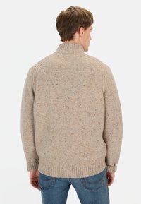 Beżowy sweter z wysokim kołnierzem, ściągaczami przy rękawach i u dołu. Nakrapiana tekstura w odcieniach brązu i niebieskiego. Noszony z niebieskimi dżinsami.