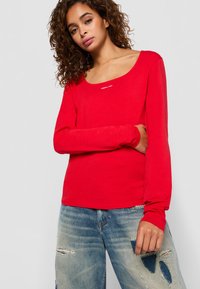 Jonge vrouw met krullend haar, gekleed in een rode lange mouwtop en versleten blauwe jeans, poseert tegen een effen lichte achtergrond.