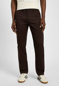 Brązowe spodnie o prostych nogawkach, wykonane z gładkiego materiału, z zamkiem błyskawicznym i zapięciem na guzik. Posiadają standardowe kieszenie i kontrastujące szwy.