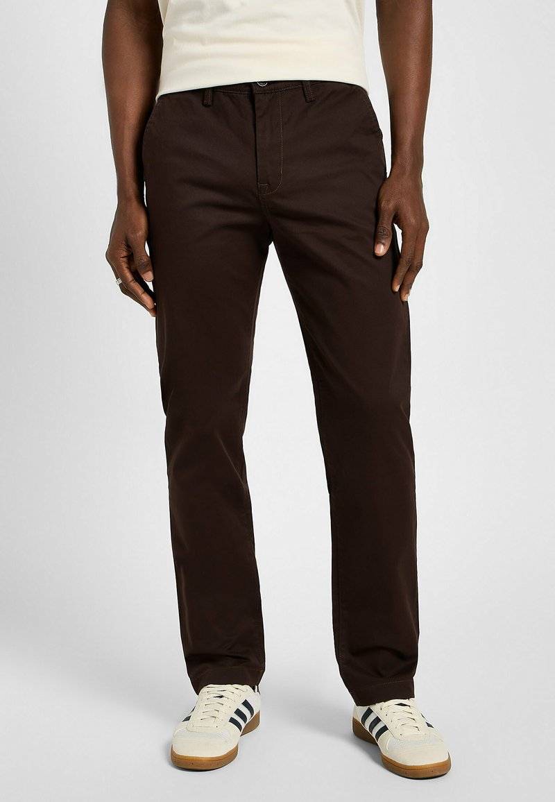 Brązowe spodnie o prostych nogawkach, wykonane z gładkiego materiału, z zamkiem błyskawicznym i zapięciem na guzik. Posiadają standardowe kieszenie i kontrastujące szwy.